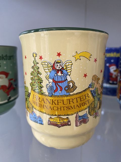 Hellgelbe Glühweintasse mit dem Dom und dem Römer versehen. Ein Engel und ein geschmückter Weihnachtsbaum halten ein Banner mit Frankfurter Weihnachtsmarkt. Darüber thront eine korpulente Frau mit Flügeln und Brezeln sowie einem Bembel in den Händen. Ein Stern mit Schweif ist ebenfalls abgebildet. 