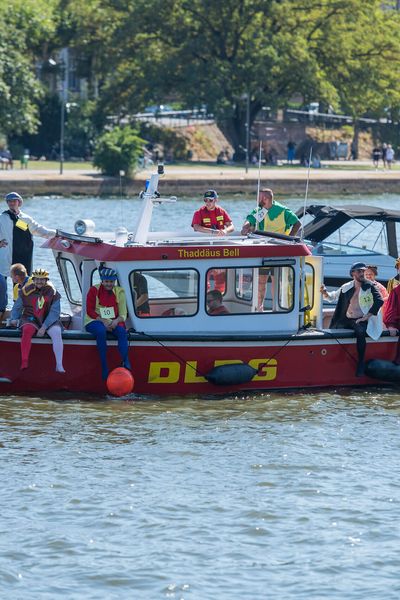 Ein DLRG-Boot mit verkleideten Teilnehmer*innen auf dem Main, sonniges Wetter und grüne Uferbäume im Hintergrund.