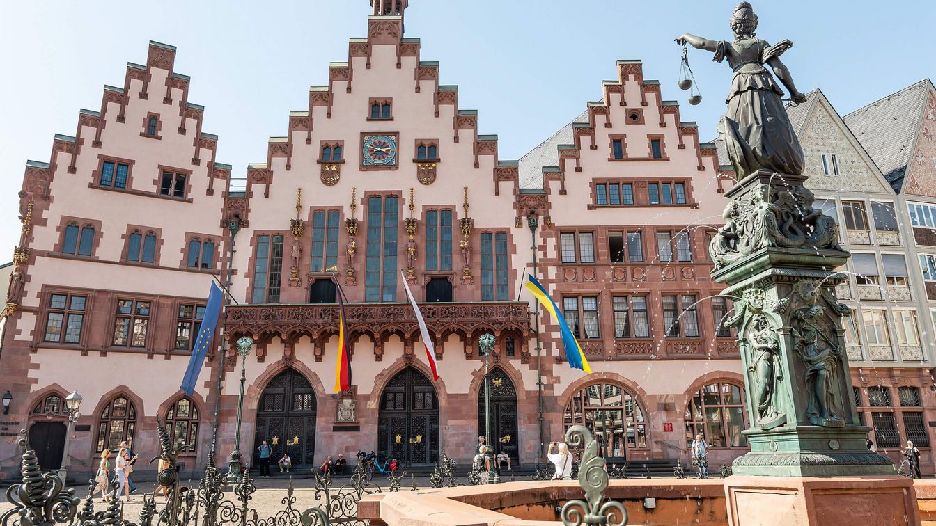 Historischer Römerberg in Frankfurt mit dem Gerechtigkeitsbrunnen im Vordergrund. Die charakteristische Fassade des Rathauses Römer mit ihren markanten Giebeln und Fahnen bildet den Hintergrund, während die Justitia-Statue auf dem Brunnen prominent hervorsticht.