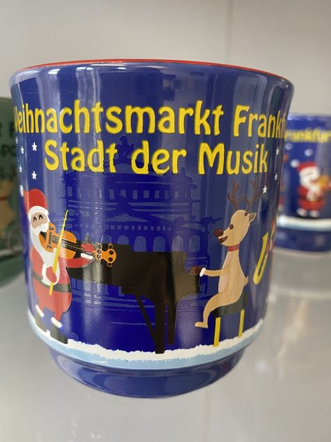 Blaue Glühweintasse mit einem Gitarren spielenden Weihnachtsmann und einem Klavier spielenden Rentier. Im Hintergrund ist die Alte Oper zu erkennen. Auf der Glühweintasse steht in gelber Schrift: Weihnachtsmarkt Frankfurt Stadt der Musik. 