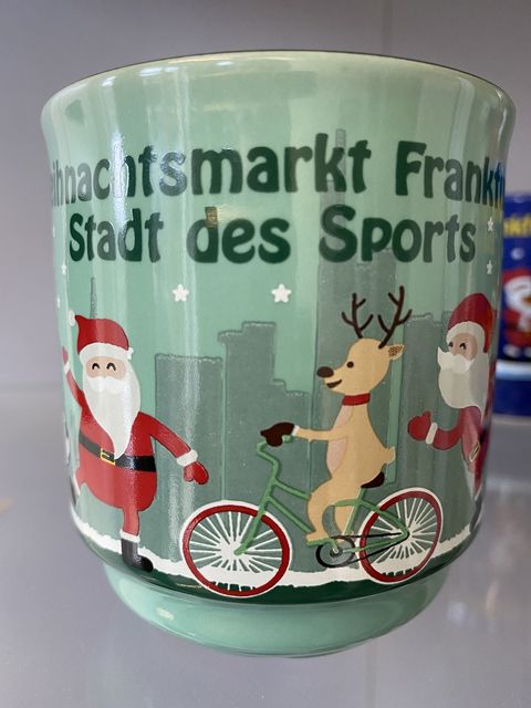 Eine Glühweintasse in mintgrün. Im Hintergrund in grau, die Frankfurter Skyline schematisch dargestellt. Im Vordergrund der Tasse ein Fahrradfahrendes Rentier, das von Weihnachtsmännern angefeuert wird. Im dunklen grün steht über dieser Szenerie Weihnachtsmarkt Frankfurt Stadt des Sports. 