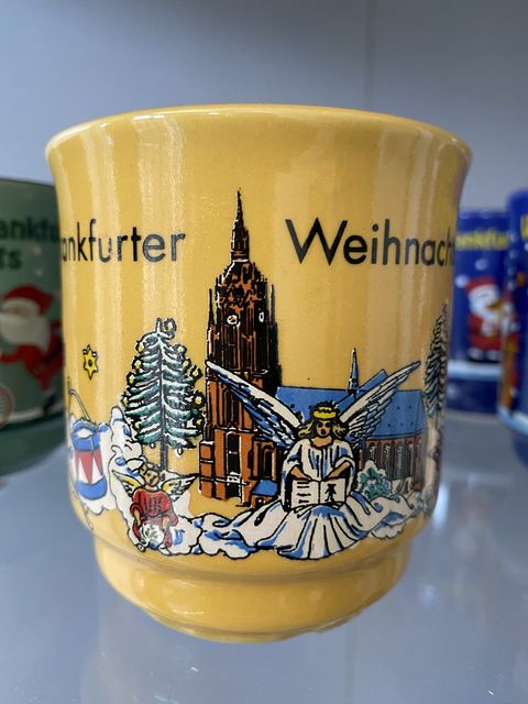 Gelbe Weihnachtsmarkttasse mit der Darstellung des Frankfurter Doms. Vor dem Dom sitzen Engel auf Wolken. Frankfurter Weihnachtsmarkt steht in schwarzer Schrift oben am Rand der Tasse. 