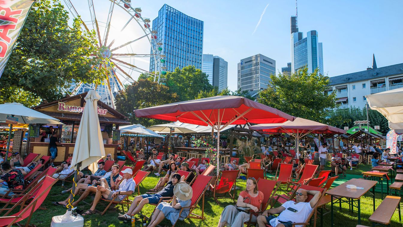 Besucher*innen entspannen in Liegestühlen unter Sonnenschirmen, mit bunten Marktständen und dem Riesenrad im Hintergrund.