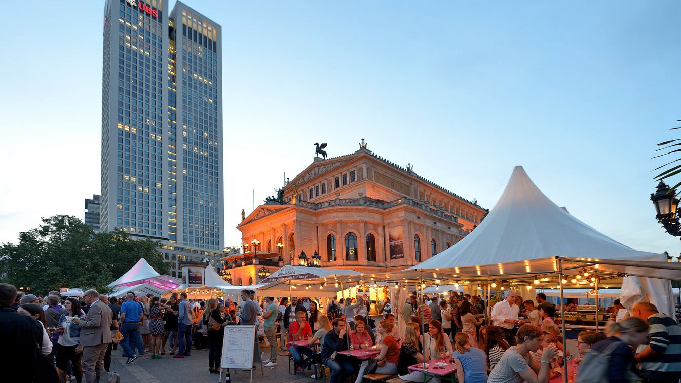 Abendstimmung auf dem Opernplatzfest in Frankfurt vor der beleuchteten Alten Oper. Besucher*innen sitzen an Tischen oder schlendern durch die Marktstände. Im Hintergrund sind moderne Hochhäuser zu sehen.