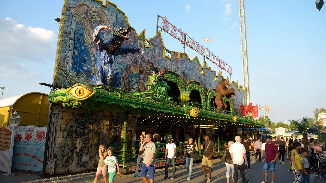 Eine Geisterbahn mit fantasievoller Fassade, gruseligen Figuren und bunten Lichtern auf einem Jahrmarkt, während Besucher*innen vorbeigehen und die Attraktion bestaunen.