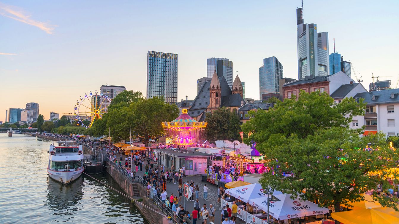 Blick auf das Mainfest mit bunten Ständen, einem Karussell, Skyline und einem beleuchteten Riesenrad bei Sonnenuntergang.