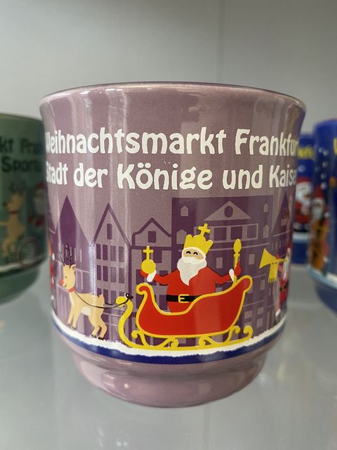 Eine Glühweintasse in helllila. Im Vordergrund eine Kutsche, die von einem Rentier gezogen wird. In der Kutsche steht ein Weihnachtsmann mit einer Krone, dem Reichsapfel in der einen Hand und dem Zepter in der anderen. Im Hintergrund ist die Frankfurter Altstadt abgebildet in einem dunklen Lila. Darüber steht Weihnachtsmarkt Frankfurt Stadt der Könige und Kaiser. 