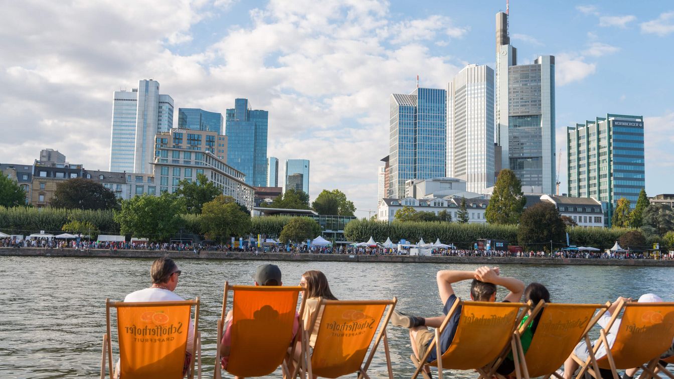 A group of friends lie in deckchairs on the banks of the Main in the sunshine and look out over the skyline.