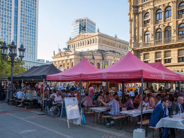 Der Rheingauer Weinmarkt in Frankfurt mit vielen Menschen unter roten Pavillons, die „Weingut der Stadt Frankfurt am Main“ zeigen. Im Hintergrund ist die Alte Oper und moderne Hochhäuser zu sehen. Eine festliche Stimmung bei gutem Wetter.