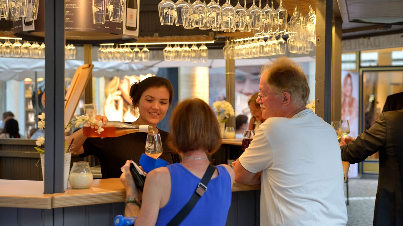 An employee pours wine at a stand while guests wait at the bar. Glasses hang around her, creating a stylish and inviting atmosphere.