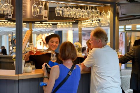 An employee pours wine at a stand while guests wait at the bar. Glasses hang around her, creating a stylish and inviting atmosphere.