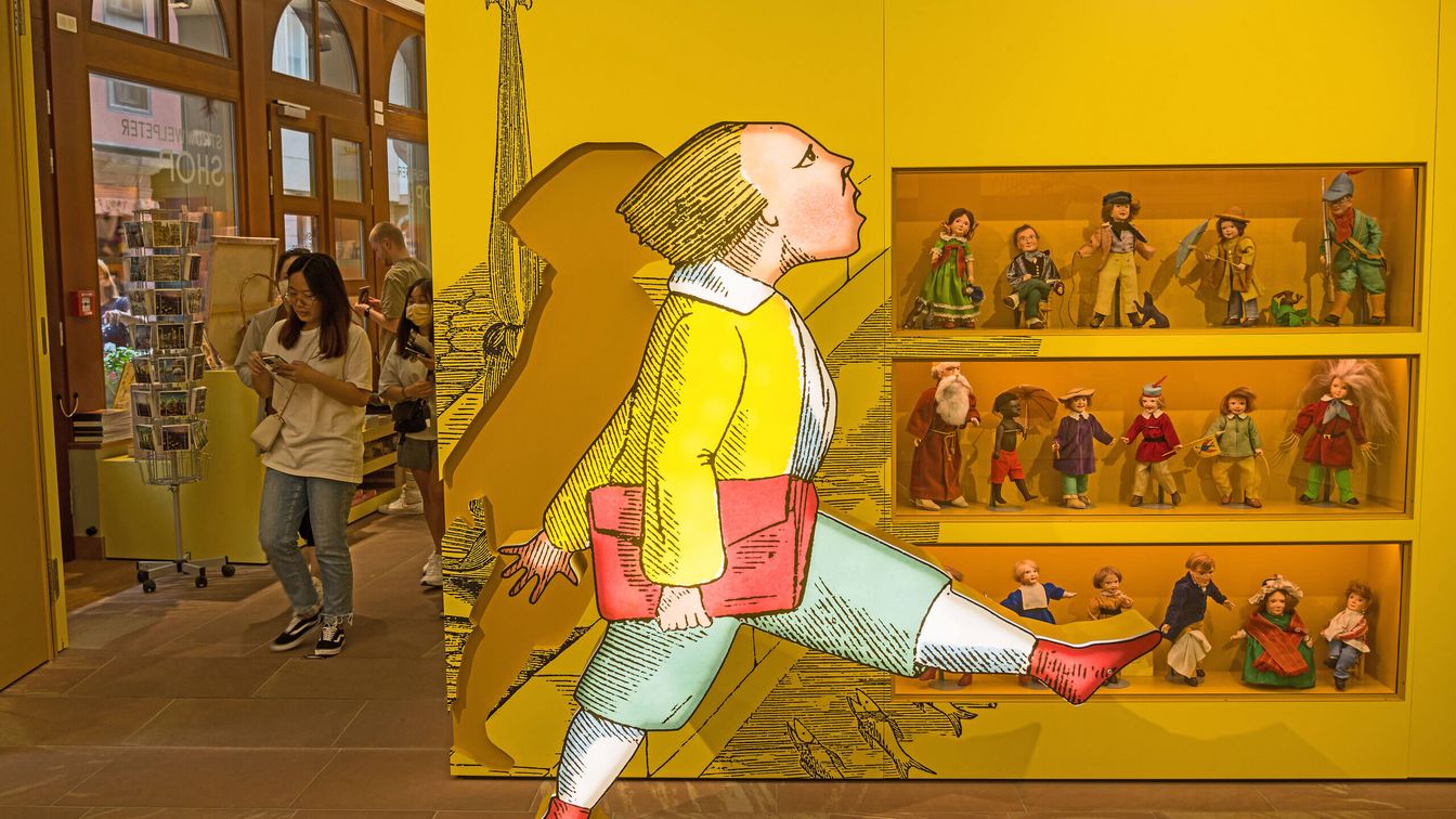 Eine Ausstellung mit einer großen Struwwelpeter-Figur, Puppen in einer Vitrine und Besucher*innen in einem gelb gestalteten Raum.