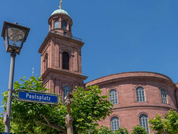 Nahaufnahme der Frankfurter Paulskirche aus rotem Sandstein mit ihrem Turm und grüner Kuppel. Im Vordergrund steht eine Laterne und ein Straßenschild mit der Aufschrift „Paulsplatz“. Der Himmel ist klar und blau.