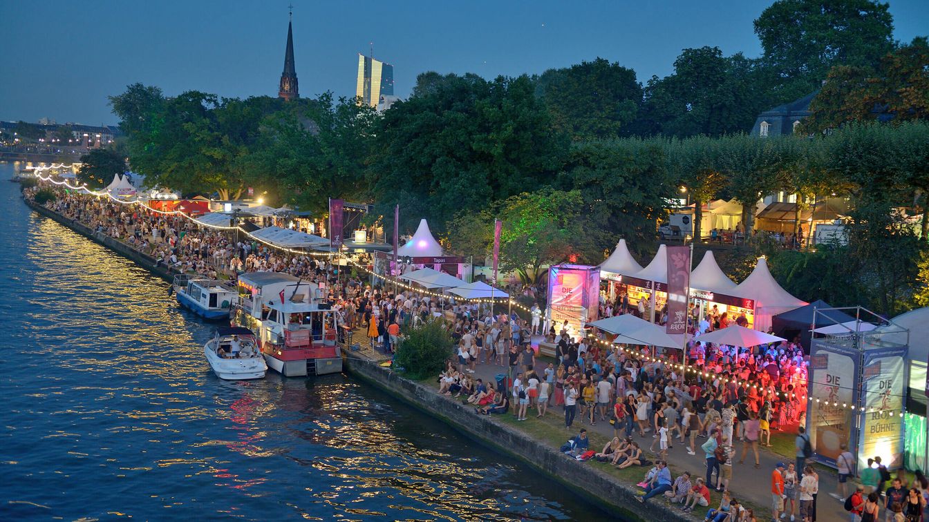 Menschenmenge entlang des Mainufers bei Dämmerung, beleuchtete Festzelte, Boote auf dem Wasser und Frankfurter Skyline im Hintergrund.