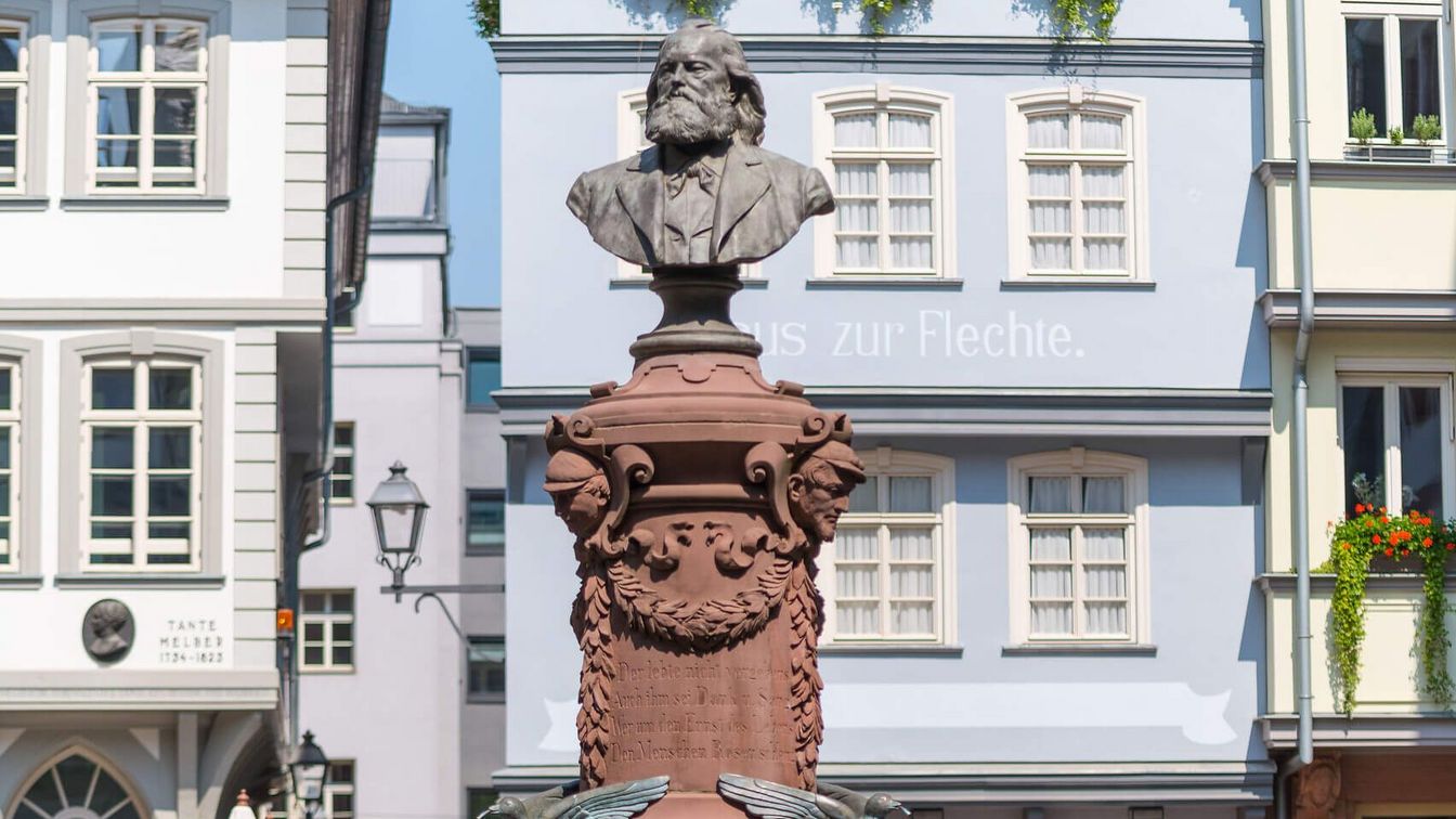 Der Friedrich-Stoltze-Brunnen in der Frankfurter Altstadt, mit einer Büste des Schriftstellers Friedrich Stoltze. Der Brunnen ist kunstvoll gestaltet und von einem schmiedeeisernen Geländer umgeben, das mit einem goldenen Adler verziert ist. Im Hintergrund sind historische Gebäude und ein kleines Straßencafé zu sehen.