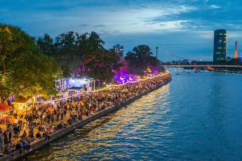 Abendliches Museumsuferfest am Mainufer in Frankfurt mit bunten Lichtern, feiernden Menschen und der beeindruckenden Skyline im Hintergrund.