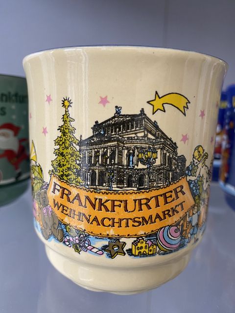 Hellgelbe Glühweintasse mit einer Darstellung der Alten Oper. Im Vordergrund ein Banner, gehalten von einem geschmückten Weihnachtsbaum und einem Engel, mit der Aufschrift Frankfurter Weihnachtsmarkt. Vor dem Banner ist ein Zimtstern, eine Zuckerstange, ein Teddybär sowie ein Kinderkreisel abgebildet. Gekrönt ist die Abbildung von einem Weihnachtsstern. 