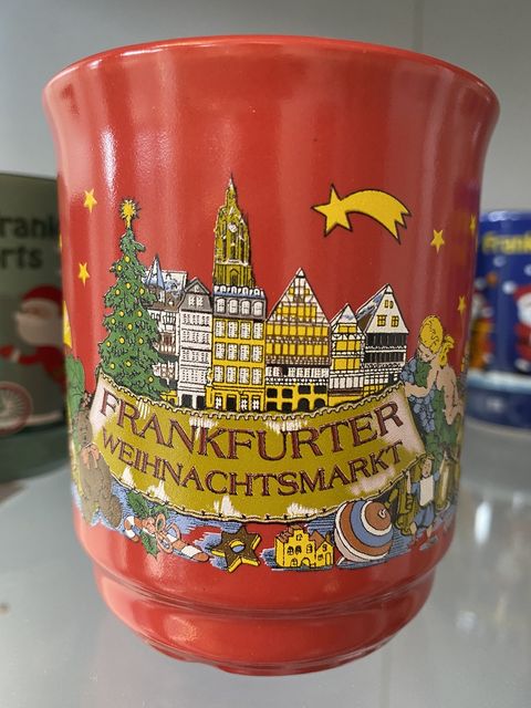 Rote Glühweintasse mit einer Darstellung des Frankfurter Römerbergs mit historischen Gebäuden. Im Vordergrund ein Banner, gehalten von einem geschmückten Weihnachtsbaum und einem Engel, mit der Aufschrift Frankfurter Weihnachtsmarkt. Vor dem Banner ist ein Zimtstern, eine Zuckerstange, ein Teddybär sowie ein Kinderkreisel abgebildet. Im Hintergrund ist der Turm des Frankfurter Doms abgebildet. Gekrönt ist die Abbildung von einem Weihnachtsstern. 