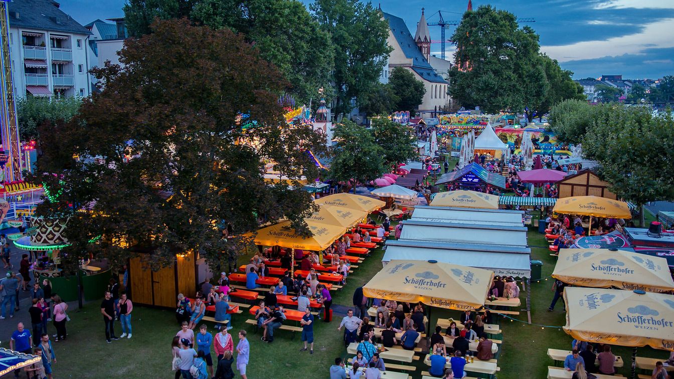 Ein belebter Biergarten mit gelben Schirmen, Holzbänken und vielen Menschen. Die Stände und Fahrgeschäfte im Hintergrund leuchten in bunten Farben.