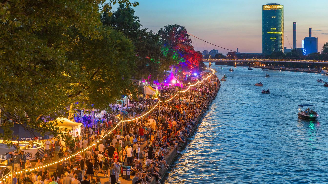 Lebhafte Festivalstimmung bei Sonnenuntergang, bunte Lichter, Besucher*innen entlang des Mainufers und Boote auf dem Wasser.