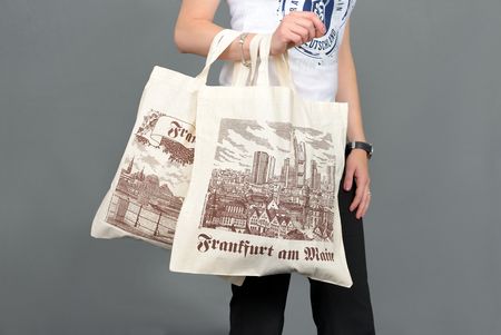 Eine Person hält zwei bedruckte Stofftaschen mit Frankfurt-Motiven. Die Taschen zeigen Skyline, historische Gebäude und den Schriftzug „Frankfurt am Main“ in braunem Druck auf beigem Stoff.