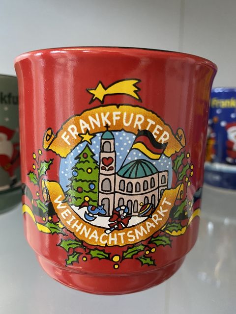 Rote Glühweintasse mit der Darstellung der Paulskirche und einem geschmückten Weihnachtsbaum in der Mitte. Über dieser Darstellung steht in weißer Schrift auf orangefarbene Hintergrund Frankfurter und darunter Weihnachtsmarkt. Um die Mitte ranken sich Blätter und gelbe Beeren. 