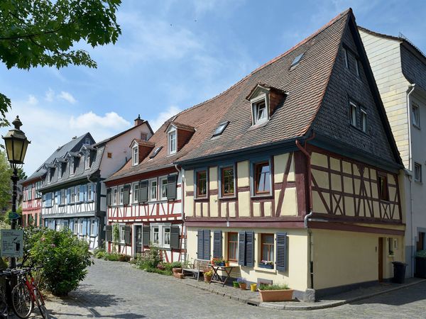 Eine malerische Fachwerkstraße in der Altstadt von Höchst mit schiefen, historischen Häusern. Die Gebäude haben bunte Fassaden, viele Fensterläden und Blumenkübel. Kopfsteinpflaster und eine Laterne prägen den ruhigen, sonnigen Straßenabschnitt.