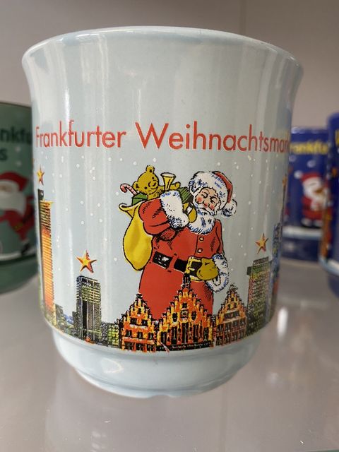 Hellblaue Glühweintasse mit der Frankfurter Skyline. Über der Skyline ist ein Weihnachtsmann mit einem Sack aus dem Spielsachen wie eine Teddybär herausstehen, zu sehen. Frankfurter Weihnachtsmarkt steht in roter Schrift darüber. 