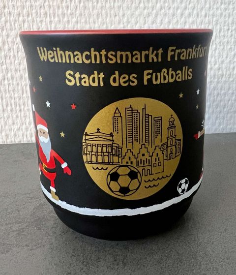 Schwarze Glühweintasse mit einem goldenen Emblem. Dort dargestellt ist eine Kirche, Hochhäuser, historische Gebäude sowie ein Fußball. Kleine Sterne in gold, weiß und rot sind im Hintergrund zu sehen. Neben dem Emblem ist ein Mann mit weißem Bart und rot/weißer Kleidung, der sich freut. Über dem goldenen Emblem steht in goldener Schrift Weihnachtsmarkt Frankfurt Stadt des Fußballs. 