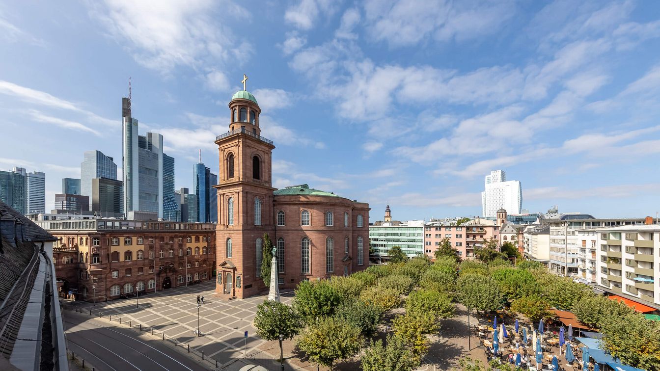 Exterior view of St Paul's Church in Frankfurt, made of red sandstone, surrounded by trees and a large square. In the background, modern skyscrapers of the Frankfurt skyline rise into the sky, which is covered by light clouds.