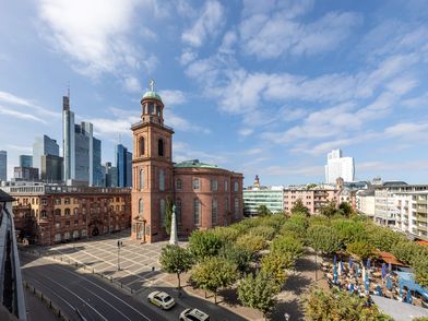 Außenansicht der Frankfurter Paulskirche aus rotem Sandstein, umgeben von Bäumen und einem großen Platz. Im Hintergrund ragen moderne Hochhäuser der Frankfurter Skyline in den Himmel, der von leichten Wolken bedeckt ist.