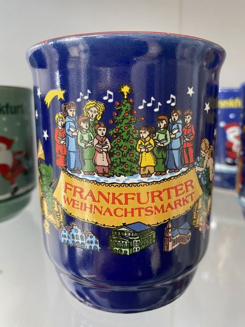 Eine blaue Glühweintasse, mit einem singenden Chor um einen festlich geschmückten Weihnachtsbaum. Auf der Tasse steht Frankfurter Weihnachtsmarkt und darunter befinden sich Abbildungen von historischen Gebäuden Frankfurts; Alte Oper, Frankfurter Dom und Roemer. 