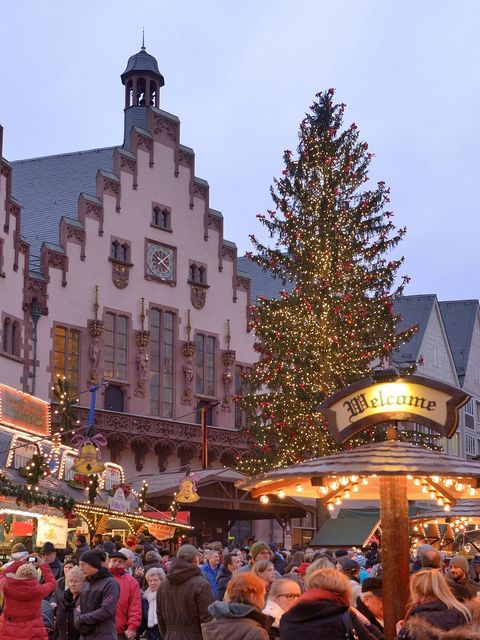 [Translate to Englisch:] Beleuchteter Weihnachtsbaum auf dem Frankfurter Weihnachtsmarkt 2014 vor der historischen Kulisse des Römers, mit geschmückten Marktständen und Menschen im Vordergrund. Das Bild zeigt eine festliche Atmosphäre in der Abenddämmerung.