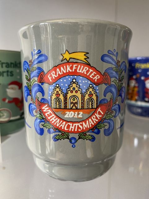 Graue Glühweintasse mit dem Frankfurter Römer als Lebkuchenhaus. Dieser ist verziert mit Zuckerstangen und Zuckerguss. Unter dem Römer steht die Jahreszahl 2012. Frankfurter Weihnachtsmarkt steht in weißer Schrift auf rotem Hintergrund um dem Römer herum. Abgeschlossen wird die Darstellung mit blauen zweigartigen Formen. 