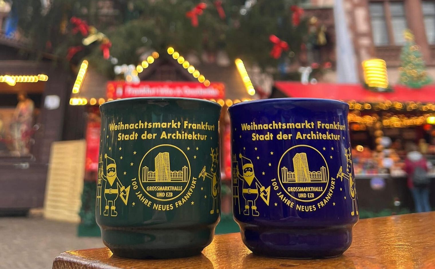 [Translate to Englisch:] Zwei Glühweintassen in grün und blau stehen nebeneinander auf einem Holztisch. Im Hintergrund ist ein geschmückter Nadelbaum zu sehen. Sowie festlich geschmückte Buden. 