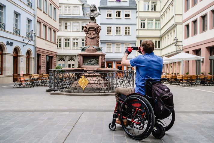 Ein Mann im Rollstuhl macht auf dem Hühnermarkt ein Foto vom Stoltze-Brunnen