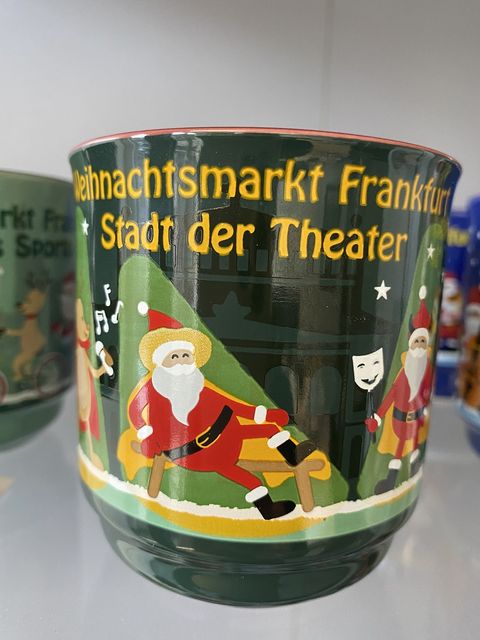 Eine grüne Glühweintasse mit Weihnachtsmännern und einem singenden Rentier. Im Hintergrund hellgrüne Bäume und das Abbild der Alten Oper. Am Tassenrand steht Frankfurter Weihnachtsmarkt Stadt der Theater. 