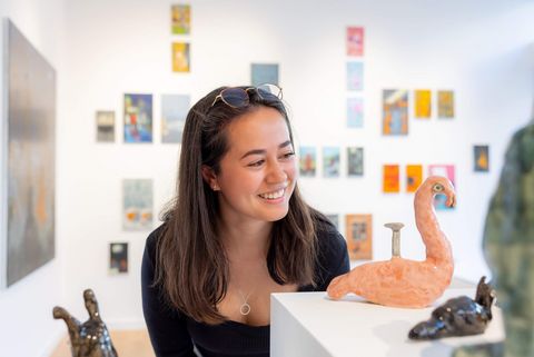Junge Frau in der Galerie, betrachtet amüsiert die Kunst, eine Schnecke aus Ton, im Hintergrund an der Wand hängen bunte kleine Bilder.