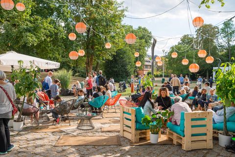 Eine lebendige Szene mit Menschen, die auf einem Festival im Freien entspannen. Bunte Möbel, Laternen und grüne Pflanzen schaffen eine gemütliche Atmosphäre unter Bäumen.