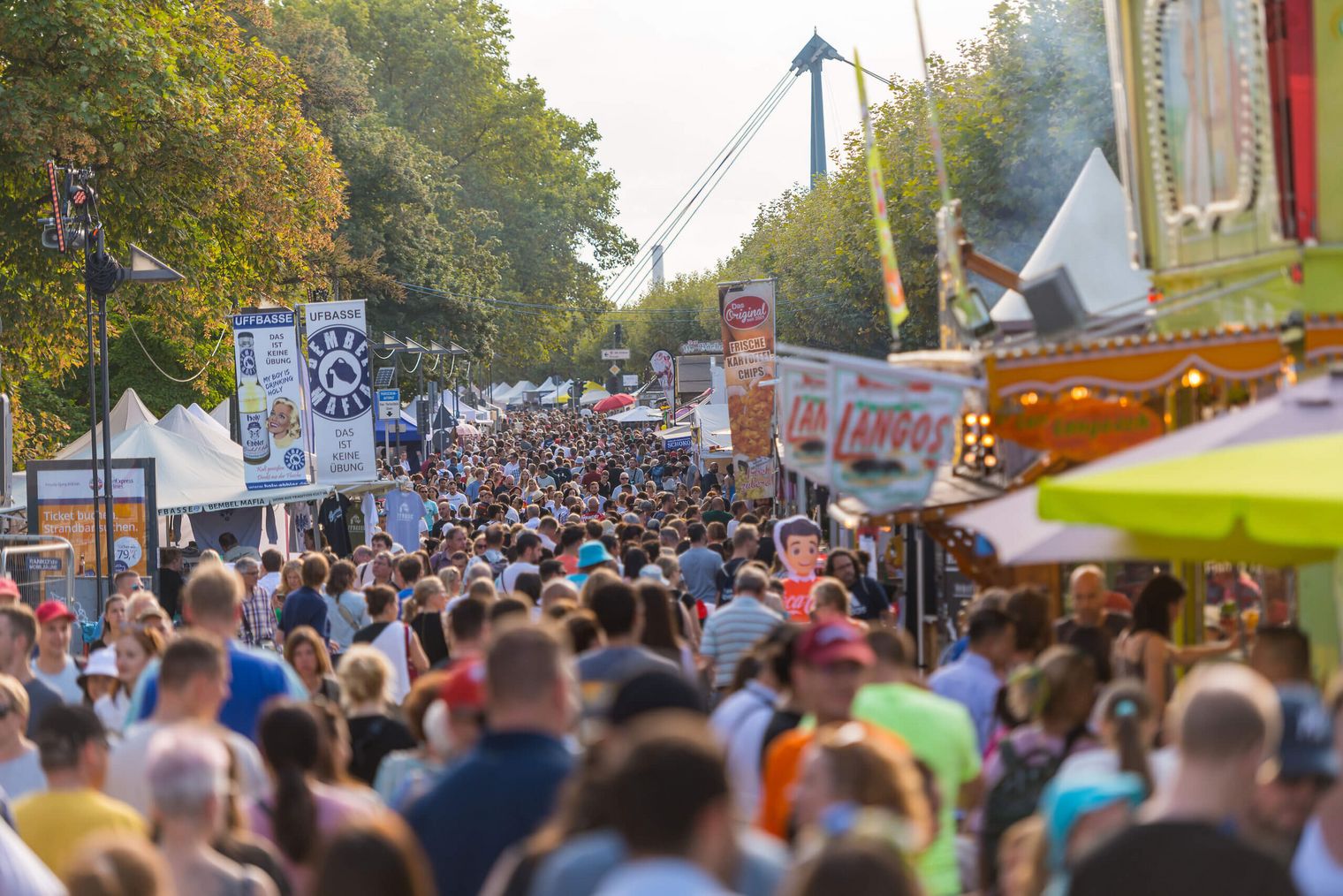 Densely packed visitors stroll through a street with food stalls and festival signs in sunny weather.