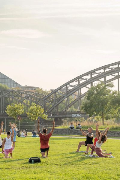 People doing yoga exercises on a meadow, in the background a bridge and a modern building.