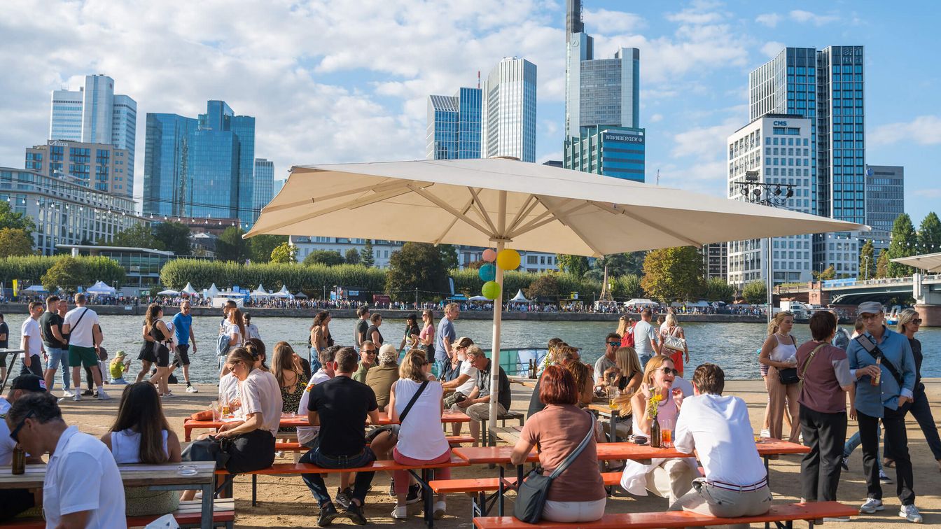 Menschen sitzen an Bierbänken unter Sonnenschirmen am Mainufer, mit Blick auf die Frankfurter Skyline und das Museumsuferfest.