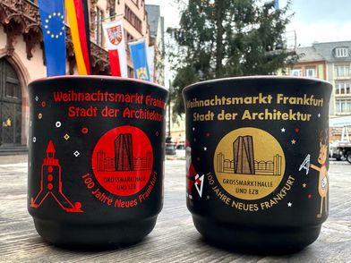 Das Bild zeigt zwei schwarze Glühweintassen vom Frankfurter Weihnachtsmarkt mit dem Schriftzug "Weihnachtsmarkt Frankfurt – Stadt der Architektur". Die linke Tasse zeigt ein rotes Motiv der Großmarkthalle und EZB, die rechte ein goldenes Motiv mit weihnachtlichen Illustrationen wie einem Weihnachtsmann und einem Rentier. Im Hintergrund sind der Römer, Flaggen und ein Weihnachtsbaum zu sehen.