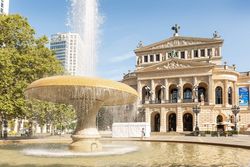 Die Alte Oper Frankfurt bei strahlendem Sonnenschein, im Vordergrund der sprudelnde Lucae-Brunnen mit Bäumen und moderner Architektur im Hintergrund.