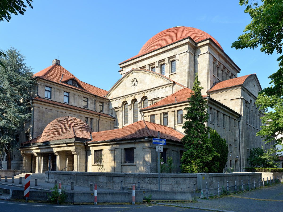 Das Bild zeigt die Westend-Synagoge in Frankfurt am Main, ein beeindruckendes jüdisches Gotteshaus mit heller Sandsteinfassade, Rundbogenfenstern und roten Ziegeldächern. Die markanten Kuppeln prägen die Silhouette, umgeben von Bäumen und einer niedrigen Steinmauer, bei strahlend blauem Himmel.