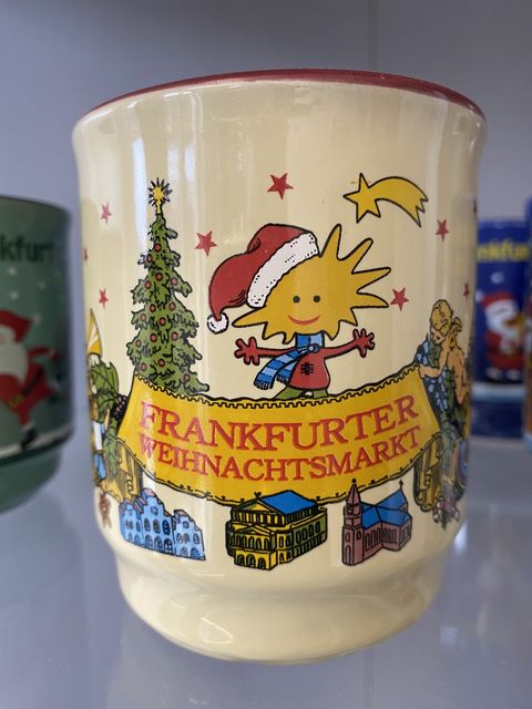 Glühweintasse in beige ist mit Frankfurts historischen Gebäuden wie der alten Oper, dem Dom und dem Römer dekoriert. Darüber ist ein Banner zu sehen, das als Schriftzug Frankfurter Weihnachtsmarkt trägt. Das Banne wird von einem Engel sowie von dem geschmückten Weihnachtsbaum gehalten. Über dem Banner ist ein weihnachtlich angezogener Stern mit Weihnachtsmütze zu sehen. Ein Stern mit Schweif komplemetiert die Darstellung. k 