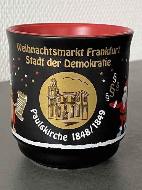 Schwarze Glühweintasse mit einem goldenen Emblem. Dort dargestellt ist die Paulskirche. Darunter steht in weißer Schrift Paulskirche 1848/1849. Neben dem Emblem ist auf der einen Seite ein Weihnachtsmann jonglierend mit Paragraphenzeichen abgebildet und auf der anderen Seite ein Rentier mit einer Schriftrolle. Kleine Sterne in gold, weiß und rot sind im Hintergrund zu sehen. Über dem Emblem steht in goldener Schrift Weihnachtsmarkt Frankfurt Stadt der Demokratie. 