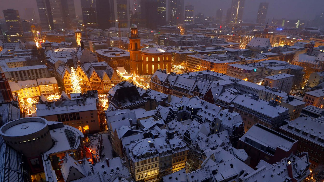 Blick über die verschneite Frankfurter Altstadt in der Abenddämmerung. Die Fachwerkhäuser und die Paulskirche sind sanft beleuchtet, während die Skyline der Hochhäuser im Hintergrund teils im Nebel verschwindet. Die warmen Lichter und schneebedeckten Dächer schaffen eine festliche, winterliche Atmosphäre.