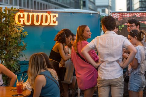 Junge Menschen unterhalten sich an einem Tisch auf einem Festival, im Hintergrund ein beleuchtetes "GUUDE"-Schild und Dekorationen.