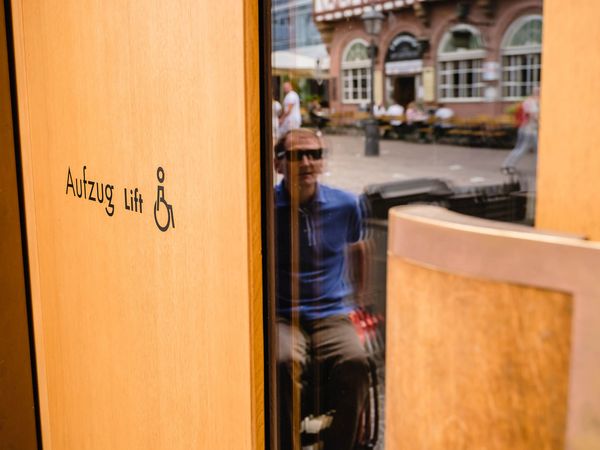 Ein Fahrstuhl mit Holzverkleidung und einem Piktogramm für Barrierefreiheit. Im Spiegel spiegelt sich ein Rollstuhlfahrer in blauem Hemd, während im Hintergrund eine belebte Altstadtstraße zu sehen ist.